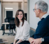 This image features two professionals engaged in a conversation in a modern office setting. The woman in the foreground is smiling confidently, while the older man listens attentively.