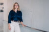 This image features a young professional woman with shoulder-length blonde hair and glasses, standing in a modern, minimalistic indoor setting. She is dressed in a navy blue blouse and white trousers, complemented by a brown leather belt.