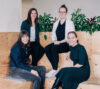 The image shows four women in a relaxed, professional setting, sitting on wooden benches. Green plants in the background create a friendly and modern atmosphere.