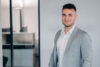 The image features a man in a light grey suit and white shirt standing in a modern office environment.